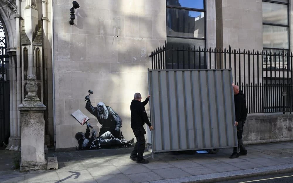 Workers installing a protective panel in front of Banksy’s mural at the Royal Courts of Justice in London. The attempt to hide the image becomes part of its history: an institutional intervention that does not erase the work, but adds another layer to its meaning. Photo: The Standard. Workers installing a protective panel in front of Banksy’s mural at the Royal Courts of Justice in London. The attempt to hide the image becomes part of its history: an institutional intervention that does not erase the work, but adds another layer to its meaning. Photo: The Standard.