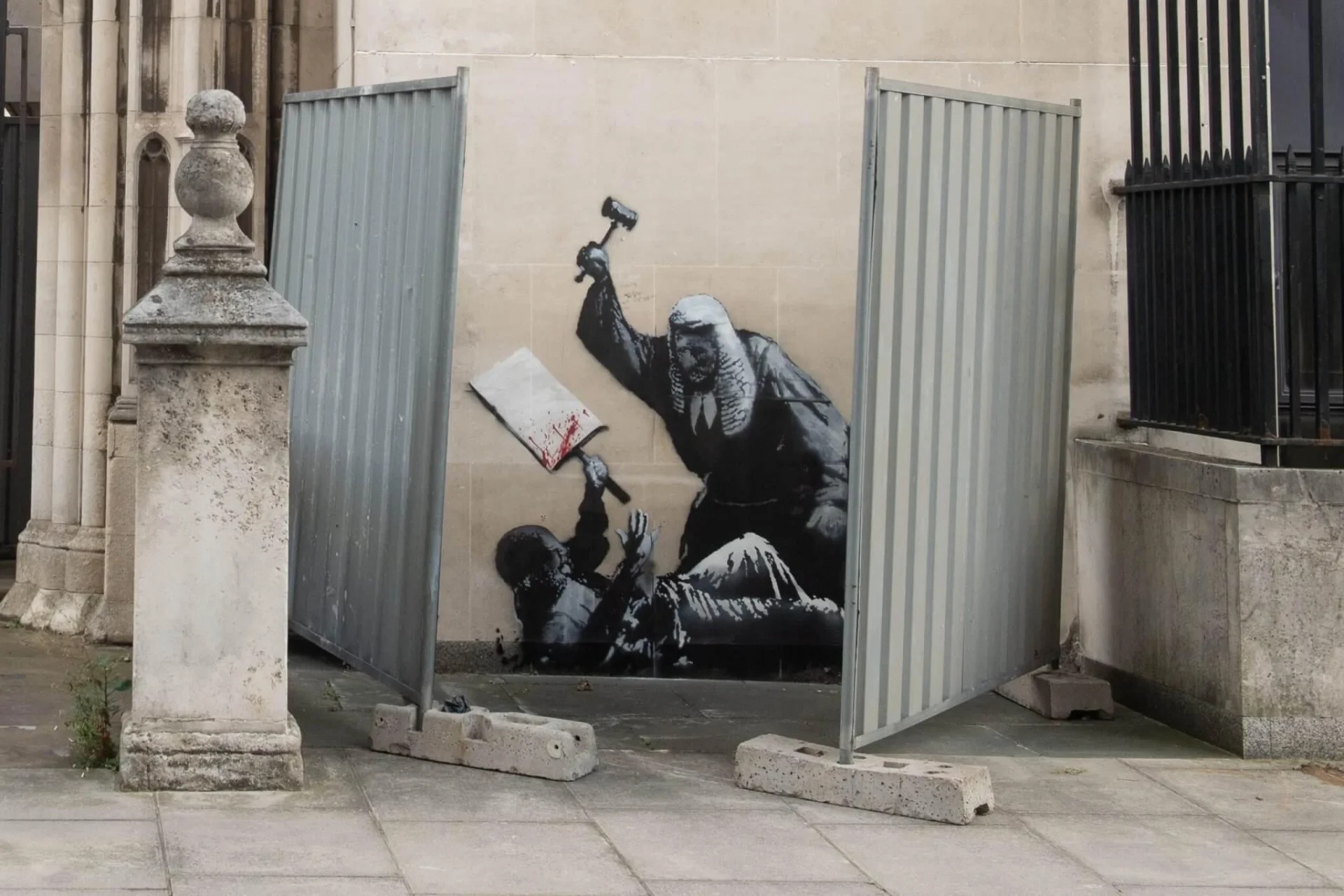 Banksy’s mural seen between two metal barriers at the Royal Courts of Justice, London. Already physically constrained by the site, the work appears almost imprisoned, making the conflict between vandalism, protection and cultural value visible before any institutional explanation begins. Photo: L’Éventail / Photonews. Banksy’s mural seen between two metal barriers at the Royal Courts of Justice, London. Already physically constrained by the site, the work appears almost imprisoned, making the conflict between vandalism, protection and cultural value visible before any institutional explanation begins. Photo: L’Éventail / Photonews.