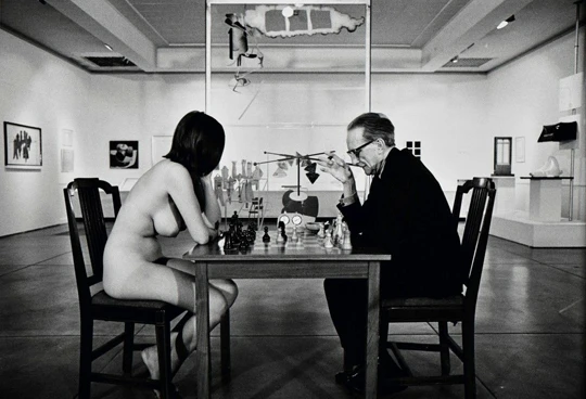 A black and white photograph showing a fully clothed elderly man, Marcel Duchamp, wearing glasses and a suit, sitting at a wooden table playing chess against a completely nude young woman, Eve Babitz. They are seated on wooden chairs facing each other in profile inside a spacious, brightly lit art gallery museum hall with other artworks visible in the background.