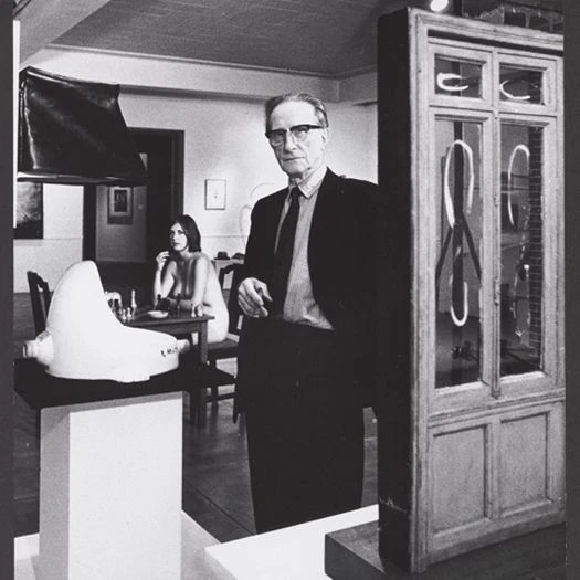 A black and white photograph featuring an elderly man, Marcel Duchamp, in a suit and glasses, looking directly at the camera. In the foreground to the left, his famous urinal sculpture "Fountain" sits on a white pedestal. To the right stands his miniature window sculpture, "Fresh Widow." In the soft-focus background, a nude young woman, Eve Babitz, is seated at a small table inside the gallery room.